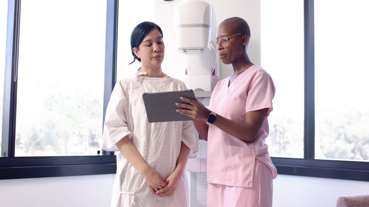 Infermiera in camice rosa che avvia una consulenza su tablet mentre la paziente ascolta in camicia da notte durante una mammografia