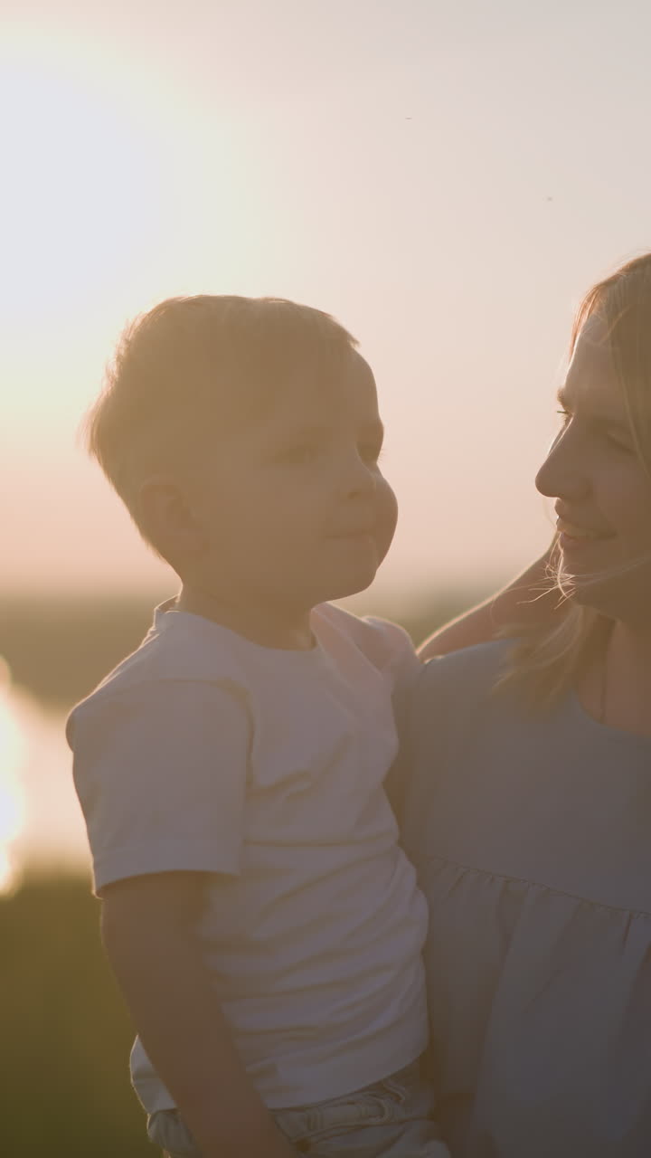 A heartwarming moment between a mother and her young son in a grassy field during sunset. The woman, dressed in a blue gown, lovingly holds her son in a white shirt as they share a tender smile