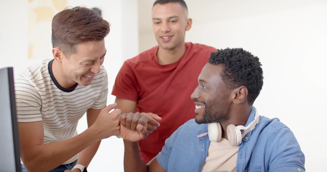 Diverse male team leaning in seeing message on smartphone and monitor, cheering at desk, copy space