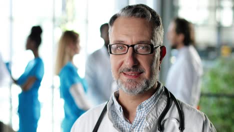 portrait of senior physician wearing a white gown, stethoscope and glasses and looking at camera and smiling in clinic