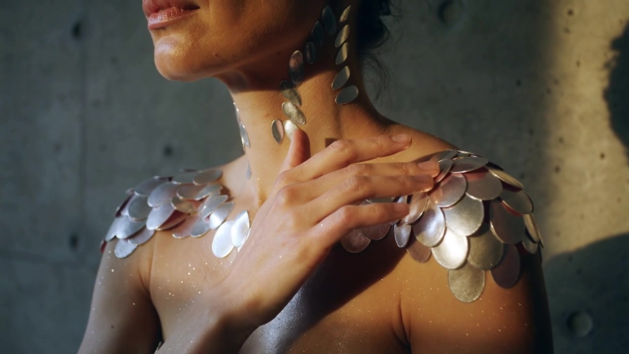 Modelo femenina deslizándose, moviendo la mano derecha hacia arriba por el pecho en un estudio, ajustando escamas metálicas.