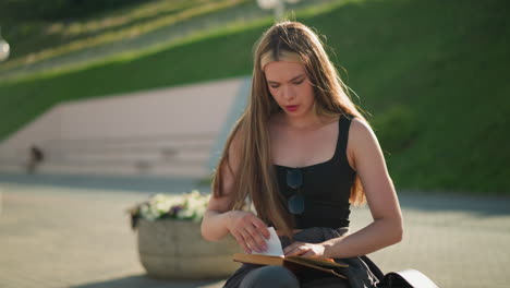 woman seated outdoors on bench with legs crossed, folding piece of paper while reading a book resting on her lap, with the sun gently illuminating her, the background features lamp poles