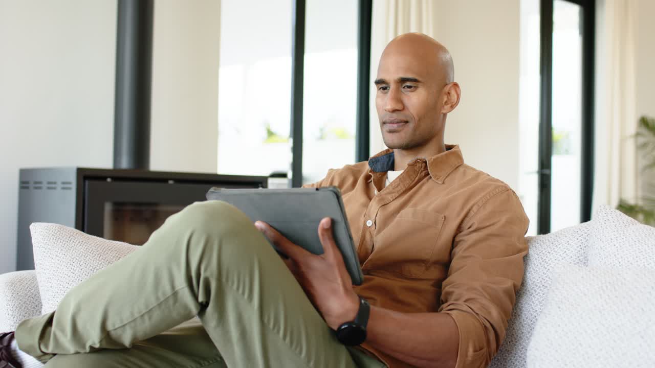 Entering living room, man in brown shirt settling on sofa and tapping tablet while reading content