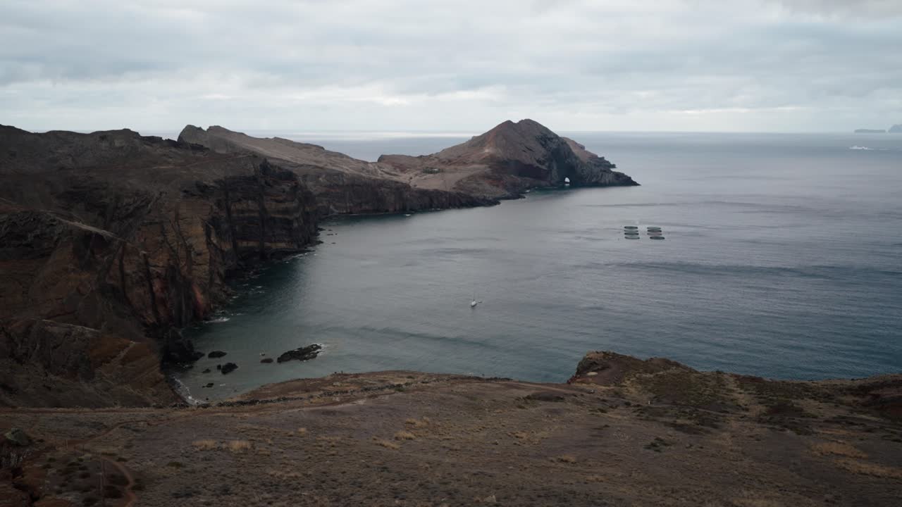 A wide aerial shot shows dramatic cliffs, a calm bay, and aquaculture pens at Ponta de São Lourenço, Madeira.