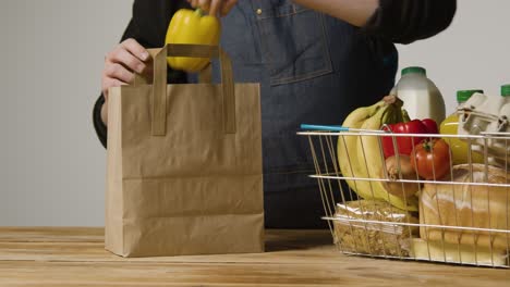 Studioaufnahme-Eines-Ladenarbeiters,-Der-Grundnahrungsmittel-Im-Supermarktdraht-Einkaufskorb-In-Eine-Papiertüte-Verpackt-4