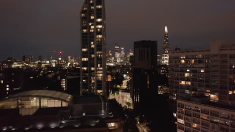 Forwards-fly-above-glowing-street-around-tall-residential-buildings.-Revealing-night-cityscape.-London,-UK