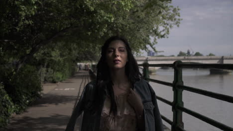 Portrait-of-an-attractive-latina-tourist-with-black-wavy-hair-and-a-jean-jacket-walking-in-a-park-in-London-with-a-view-of-Putney-bridge-behind-her