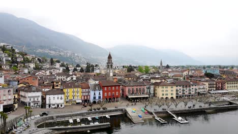 Vista-Aérea-De-Ascona-En-Ticino,-Suiza-En-La-Orilla-Del-Lago-Maggiore-Con-Un-Movimiento-De-Alejamiento-Que-Revela-Los-Tejados,-El-Lago-Y-Las-Montañas-En-El-Fondo-Desde-Una-Elevación-Más-Alta