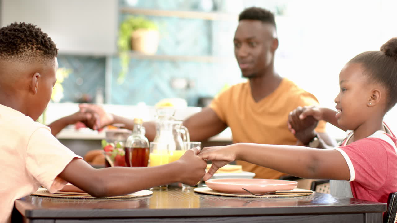Famiglia afroamericana che si tiene per mano e prega prima di pranzo, seduta al tavolo da pranzo
