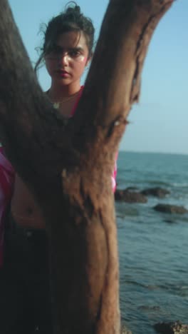 young woman standing by the beach, posing behind a tree with ocean view