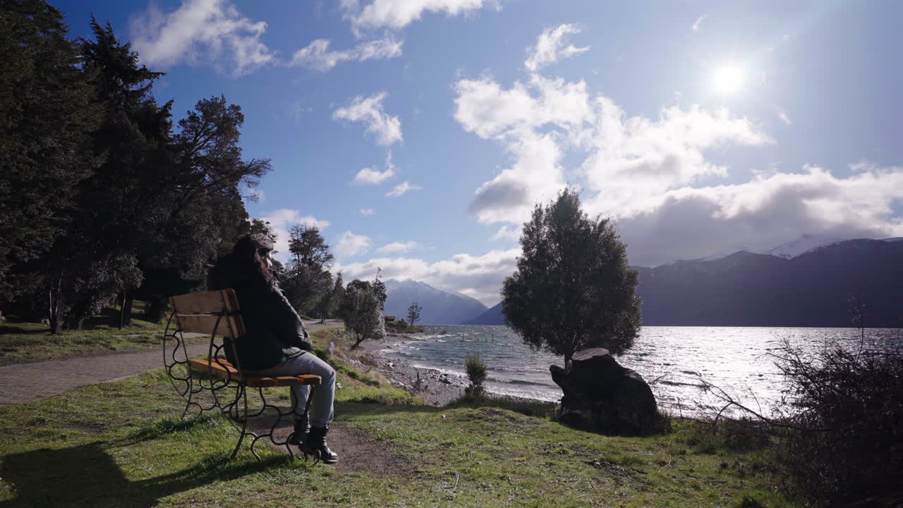 Woman sits alone on a bench overlooking the windswept waters of Villa Traful, Argentina, surrounded by trees and mountains under a bright sun and scattered clouds wearing winter or autumn garment