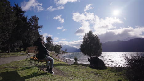 woman sits alone on a bench overlooking the windswept waters of villa traful, argentina, surrounded by trees and mountains under a bright sun and scattered clouds wearing winter or autumn garment