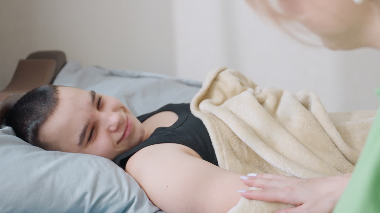Close up of boy sleeping peacefully under blanket as caring mother gently touches his nose with hand making him wake up slowly and smile warmly in cozy bedroom filled with morning tenderness