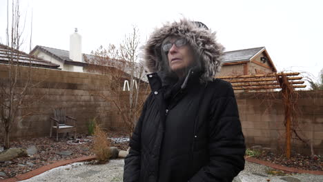 una anciana con un grueso abrigo de invierno y una capucha parada en una helada tormenta de nieve de invierno a cámara lenta