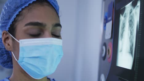 Mixed-race-female-surgeon-wearing-protective-clothing-looking-at-x-ray-on-screen