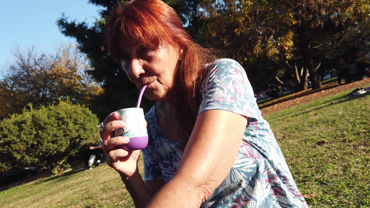 Red haired Caucasian senior woman drinks mate in an urban outdoors green park, portrait view, South American beverage