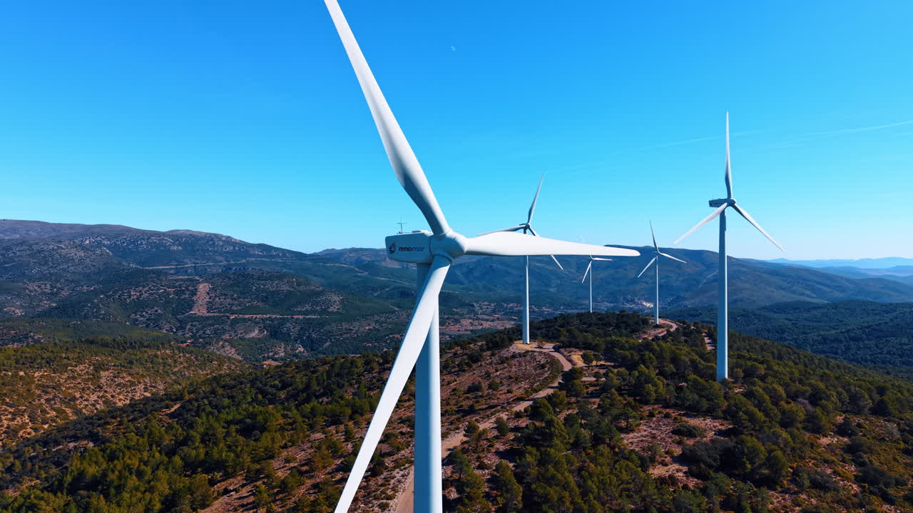 Madri, Espanha, 8 de agosto de 2025: Vista de baixo ângulo das pás de uma turbina eólica sob a luz do sol. Perspectiva para cima de pás brancas de turbinas eólicas girando sobre colinas arborizadas e cadeias de montanhas distantes