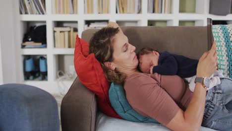 video de una madre caucásica con una tableta acostada en el sofá con un bebé recién nacido durmiendo