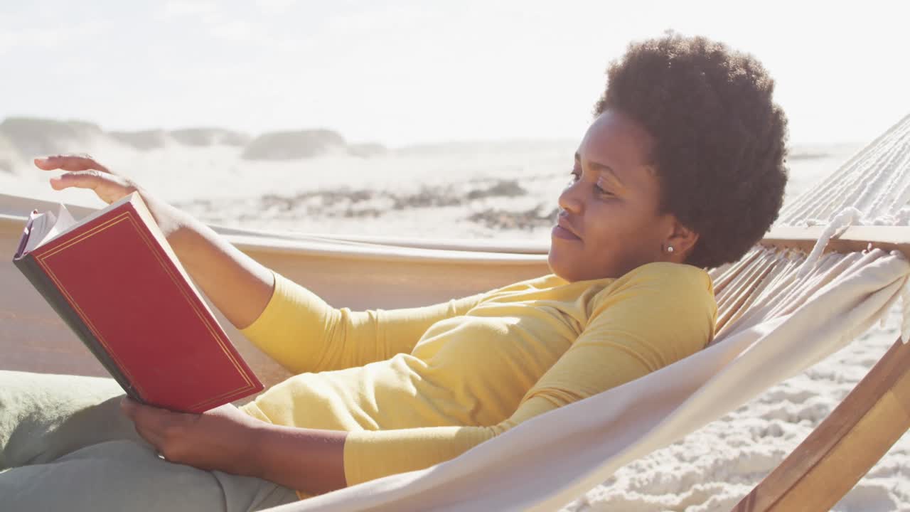 Glad afroamerikansk kvinde, der læser og ligger i hængekøje på solrig strand