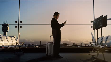 full body side view of asian businessman with rolling suitcase in boarding lounge of airline hub, using a tablet while waiting for flight, airport terminal with airplane takes off outside the window