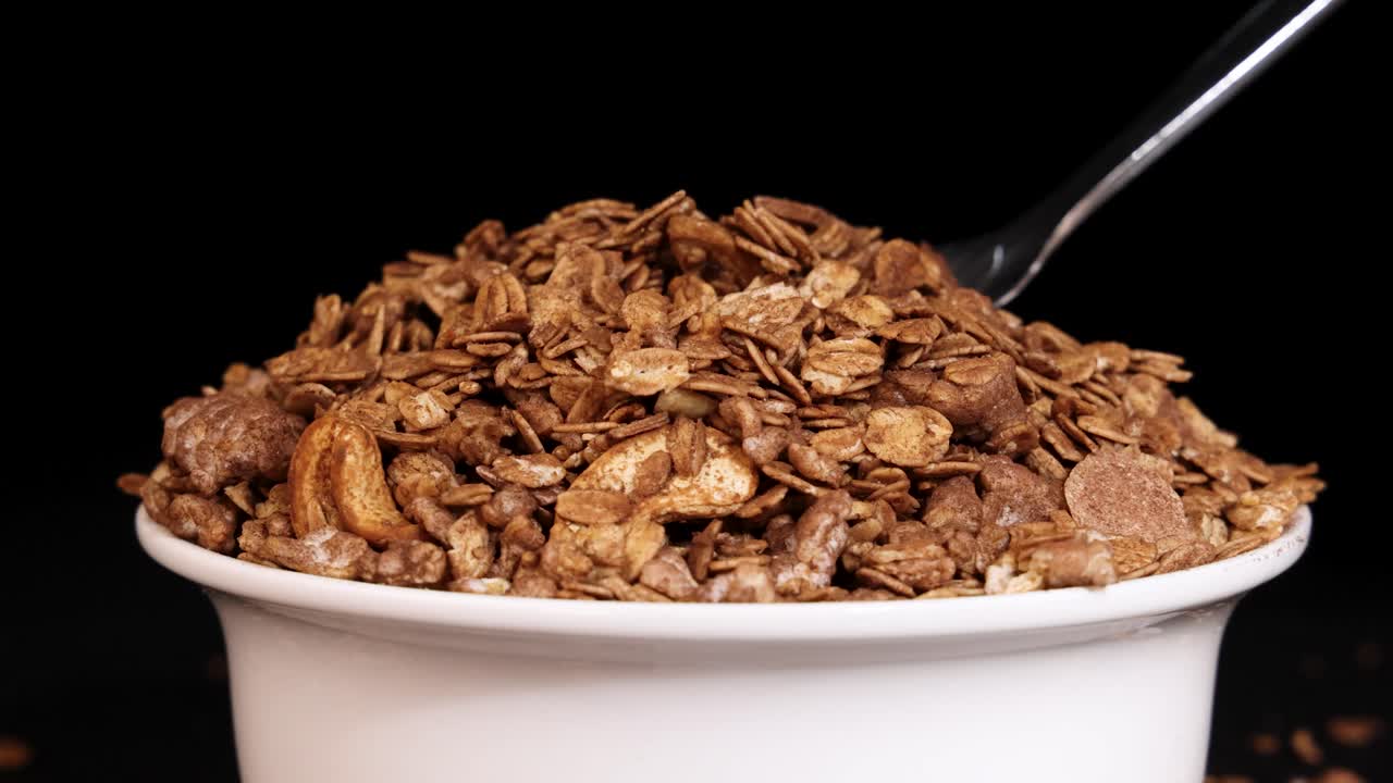 A spoon drops granola into a bowl against a black background, highlighting texture and motion in a close-up view