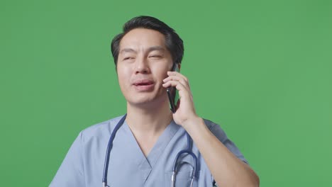 병원 에서 녹색 스크린 배경 에 서 있는 동안 스테토스코프 를 들고 스마트폰 으로 대화 하는 아시아인 남성 의사 의 근접 사진