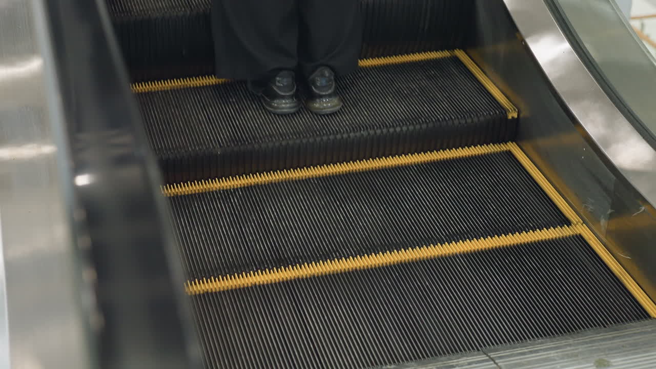 Close up rear view of person stepping onto ascending escalator in brightly lit shopping mall, showing motion of steps with yellow safety lines, metallic edges