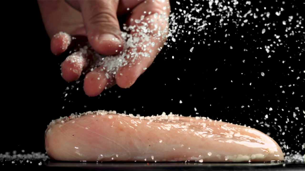 Hand Sprinkling Salt on Raw Chicken Breast in Slow Motion