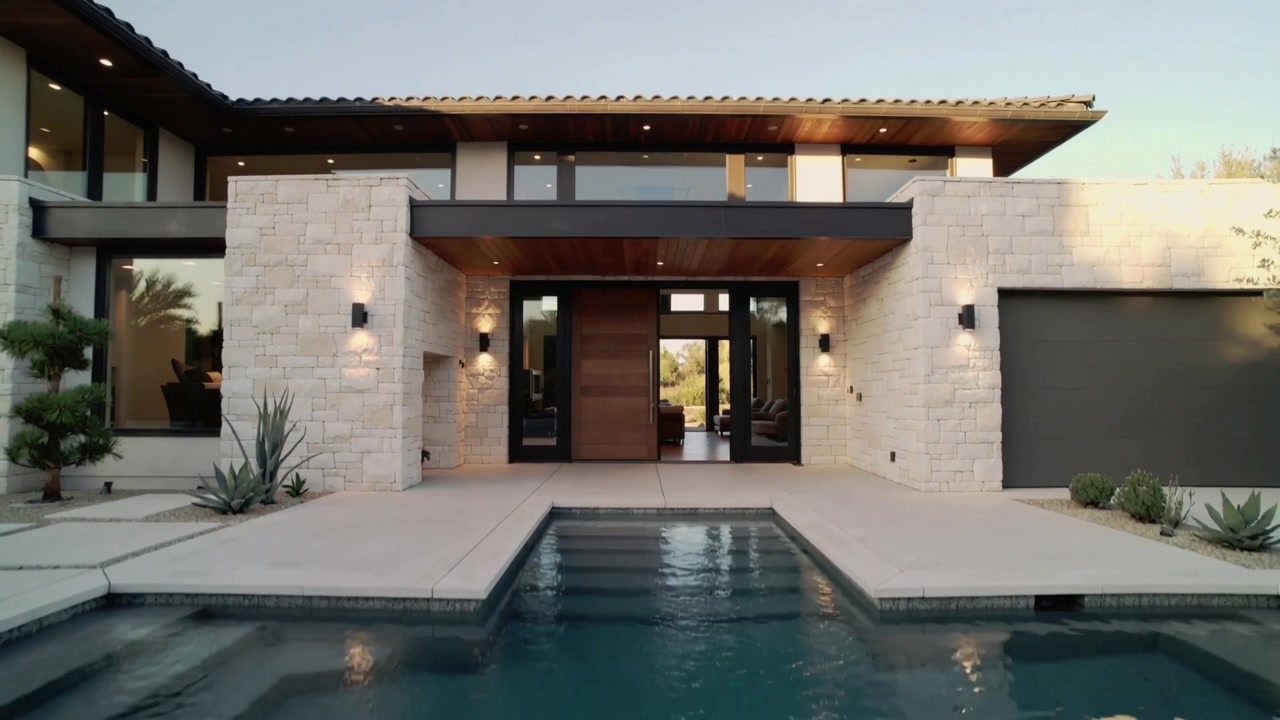 La telecamera si allontana rivelando la facciata di una casa moderna e una piscina a sfioro all'ingresso principale, mostrando luce.