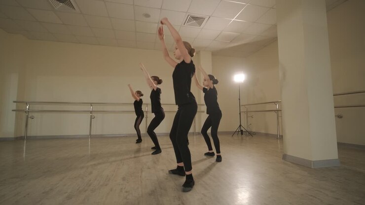 Un gruppo di giovani allievi di danza classica in abbigliamento da ballo nero che eseguono esercizi in un ampio studio di danza con pavimenti in legno e barre a muro. Espressioni concentrate e movimenti sincronizzati