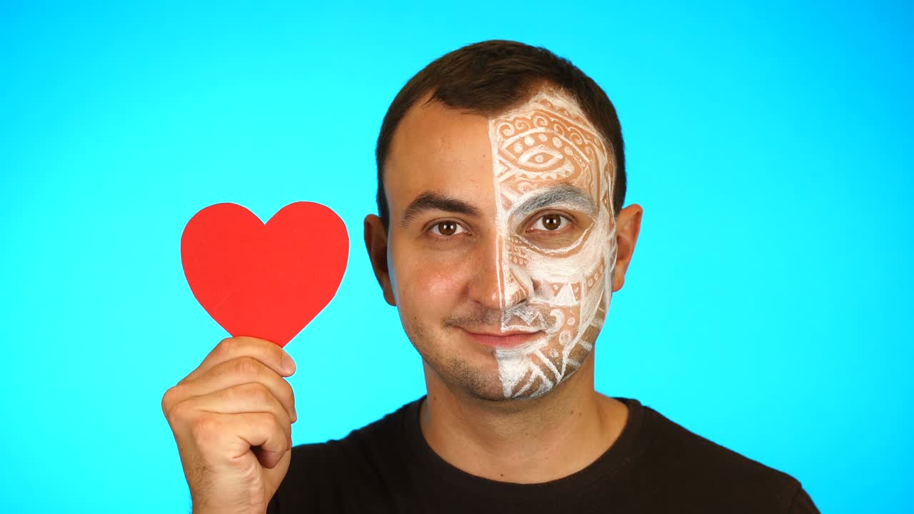 Uomo con il viso dipinto tiene in mano un cuore di carta rosso e guarda la telecamera su sfondo blu.