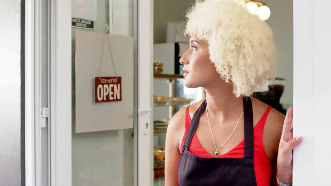 Young biracial woman with curly blonde hair stands by an open cafe door