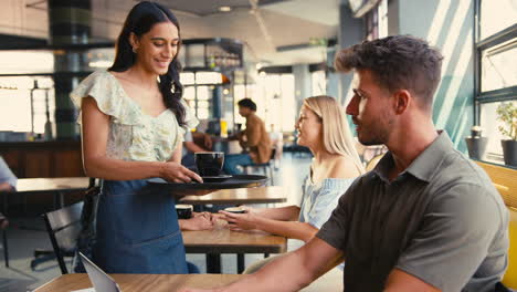 waitress serving drink to man working on laptop in busy coffee shop