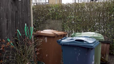 Leichter-Schnee-Fällt-Auf-Müll--Und-Recyclingbehälter-Im-Vorgarten-Neben-Der-Hecke