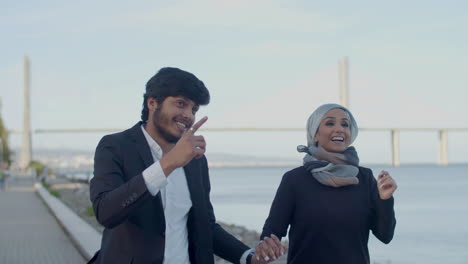 cheerful arabic lovers running next to river and holding hands