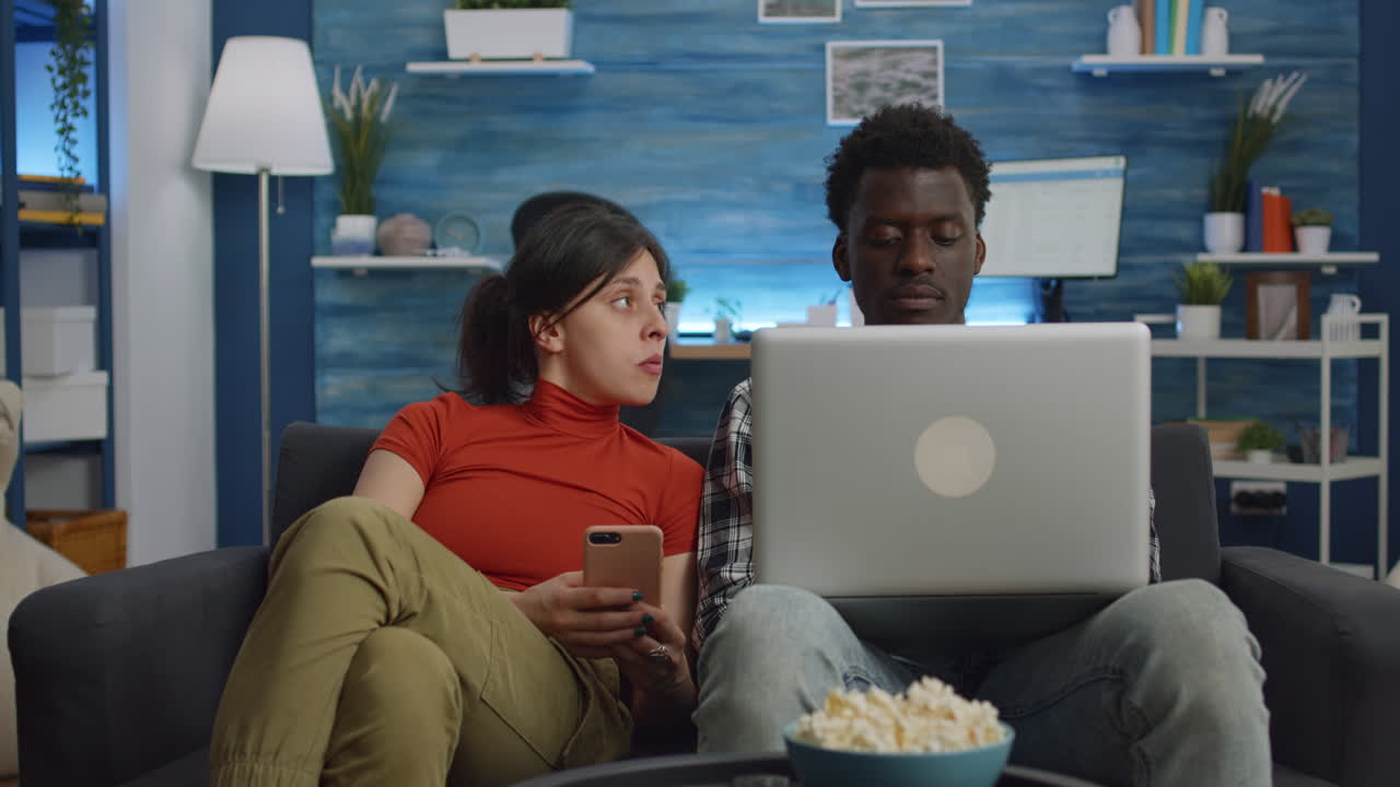 Modern interracial couple with technology sitting on sofa