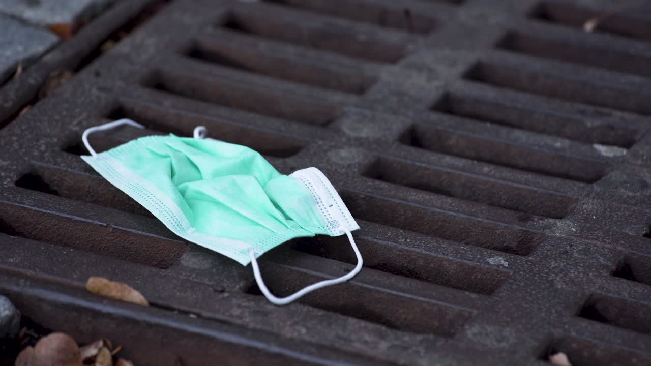 Una semplice mascherina protettiva verde per il Covid-19 abbandonata su un tombino arrugginito tra foglie secche, una persona che ci cammina sopra graffiandola con la punta della scarpa, ripresa statica ravvicinata in 4K