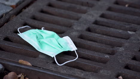 a simple green protective covid-19 face mask lying thrown out on a rusty sewerage hatch amongst dry leaves, a person walking over it, scratching it with the tip of his shoe, static close up 4k shot