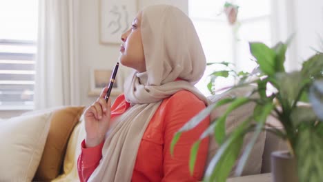 Video-of-thoughtful-biracial-woman-in-hijab-holding-pen-and-writing-in-living-room-at-home