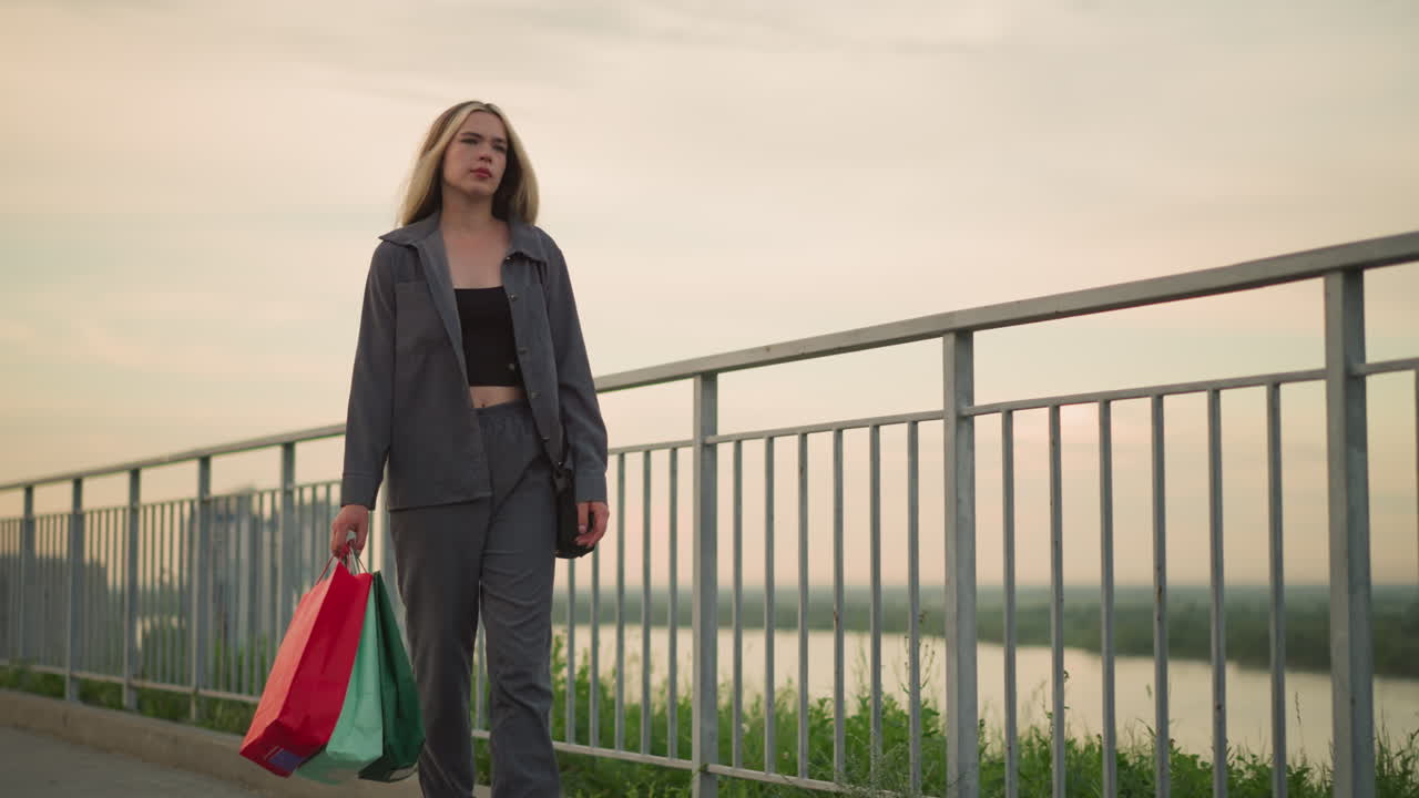 회색 옷을 입은 여자가 레일을 따라 걸어가고, 다채로운 쇼핑 가방과 검은 드백을 들고, 그녀는 피곤해 보이며, 배경에는 강을 가로지르는 먼 다리의 흐릿한 시야가 있습니다.