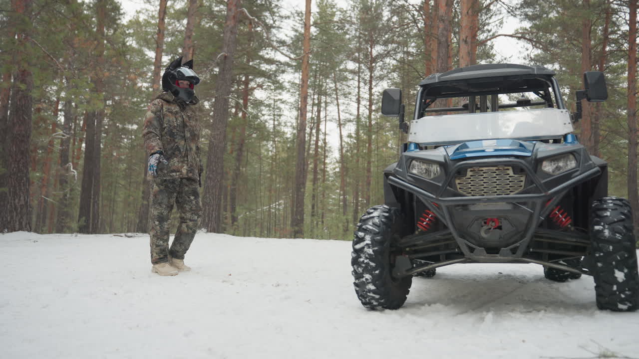 Pilota mimetizzato accanto all'ATV nella neve, in un ambiente di pineta, con il robusto veicolo fuoristrada parcheggiato; il pilota ispeziona il verricello e la griglia anteriore, indossando tuta mimetica completa e casco, con pneumatici coperti di neve, sotto una luce nuvolosa e in un'atmosfera silenziosa.