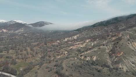 A-drone-shot-of-forest-mountains-with-snow,-warm-weather,-little-village,-flying-towards-a-foggy-hill,-4K-video,-European-winter-nature,-scenic-white-clouds,-beautiful-landscape