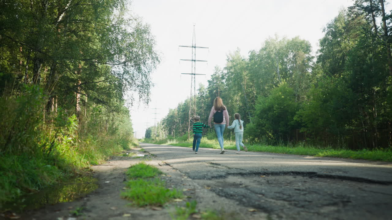 Back view of woman holding hands with two children as they run together on quiet tarred road through lush forest area, surrounded by greenery and sunlight