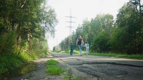 back view of woman holding hands with two children as they run together on quiet tarred road through lush forest area, surrounded by greenery and sunlight