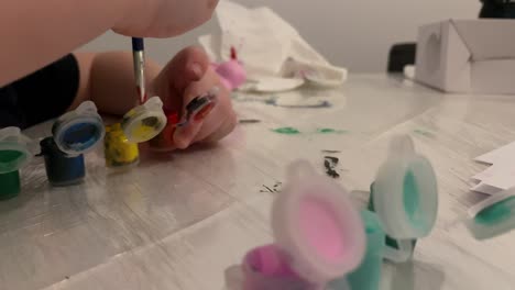 the child mixes with a paintbrush in the paint container. coloured paint containers on the table. the child is playing at home. close-up