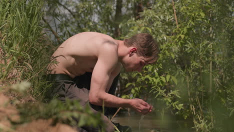 soldier crouched beside river shore with one knee grounded and elbow resting on thigh, torso smeared with mud, camouflage face paint, peaceful waters and lush foliage swaying in daylight breeze
