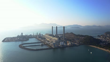 Drone-shot,-traveling-to-the-left,-of-a-big-coastal-coal-fired-power-plant-on-an-island-next-to-a-village-with-a-beach-during-a-beautiful-sunny-day-1