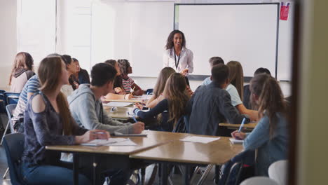maestra de secundaria haciendo preguntas de pie junto a una pizarra interactiva enseñando una lección