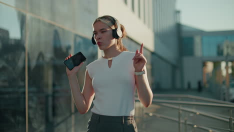 young girl in white sleeveless top dancing energetically while holding smartphone and wearing headphones, enjoying music outdoors with sunlight reflection and residential building