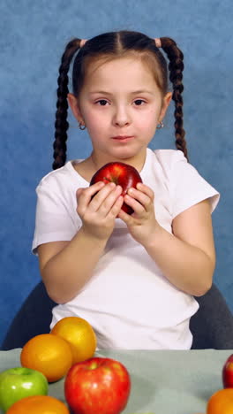 little girl bites red apple on blue background. cute child eating a fresh fruit near the table full of different organic fruits. healthy food. vertical video